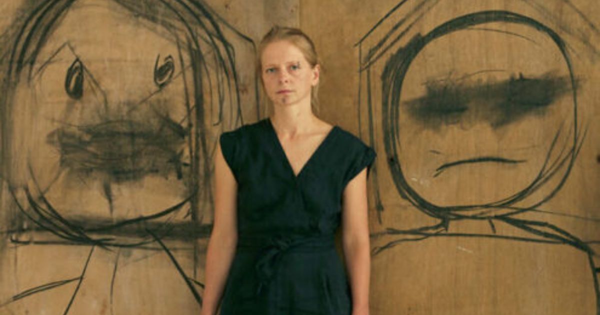 Alt text: A person stands in front of a brown wooden wall with two large, rough black charcoal-style drawings on either side. Each drawing shows an abstract face with exaggerated circular eyes and simple mouth shapes. The person, dressed in dark clothing, is centred between the two expressive, childlike sketches.