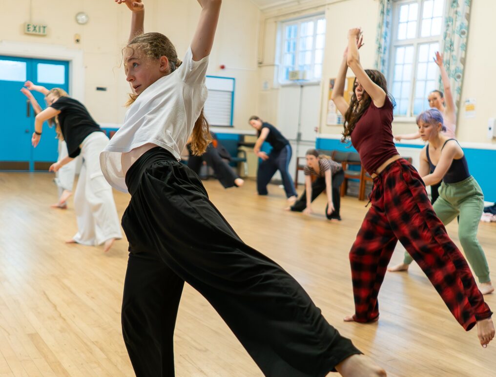 A group of people practice a dance routine in a studio with wooden floors, large windows, and bright lighting.
