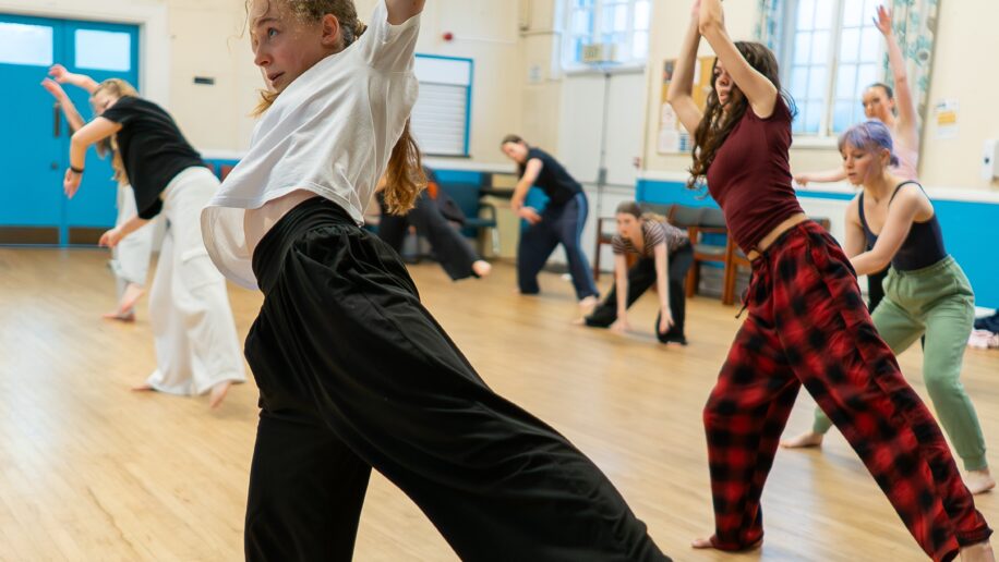 A group of people practice a dance routine in a studio with wooden floors, large windows, and bright lighting.