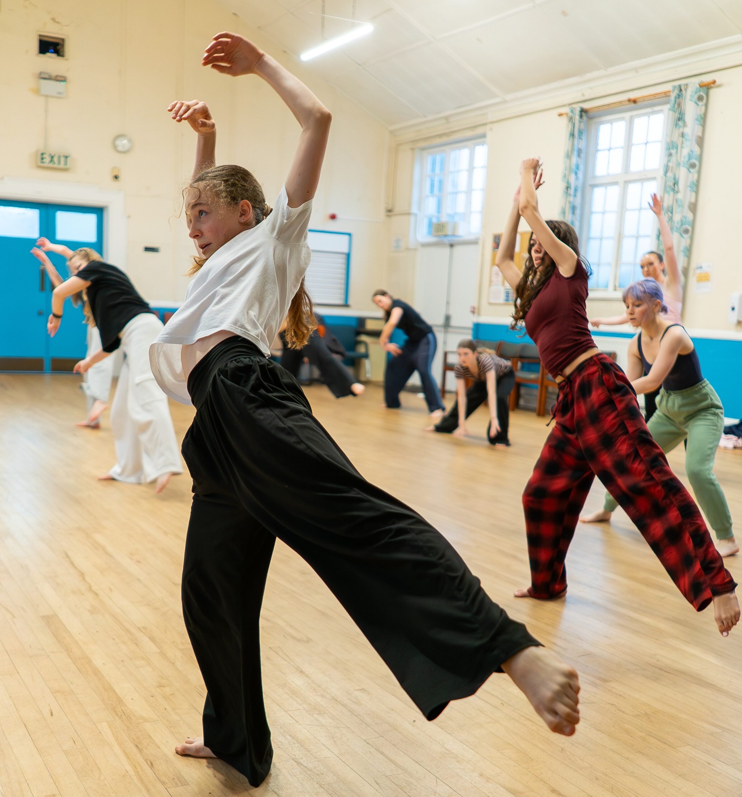 A group of people practice a dance routine in a studio with wooden floors, large windows, and bright lighting.