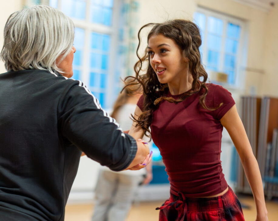 A young woman in red plaid pants dances energetically with an older woman in a bright room. They are smiling, conveying joy and connection.