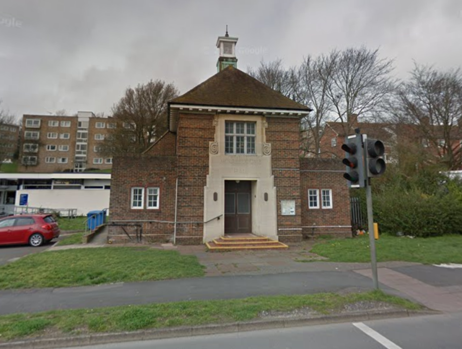 A quaint, small brick building with a steep, tiled roof and central cupola, sits by a road with traffic lights. It conveys a historic, serene charm.