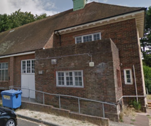 A brick building with a triangular roof and green cupola is surrounded by greenery. A black car and blue recycling bin are in front under a cloudy sky.