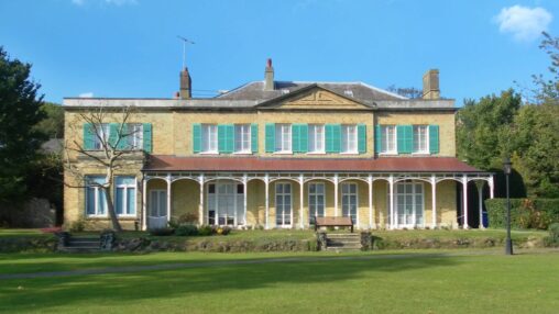 Historic two-story house with green shutters and arches, set in a lush, expansive lawn under a blue sky, evokes a serene and timeless elegance.