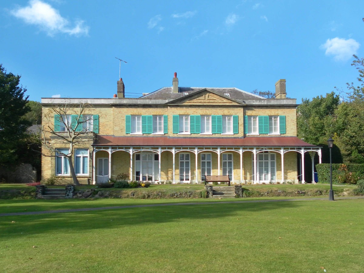 Historic two-story house with green shutters and arches, set in a lush, expansive lawn under a blue sky, evokes a serene and timeless elegance.