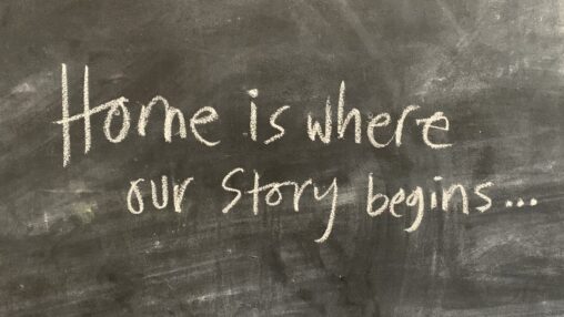 Chalkboard background with handwritten text in white chalk reads "Home is where our story begins..." conveying warmth and a sense of beginning.