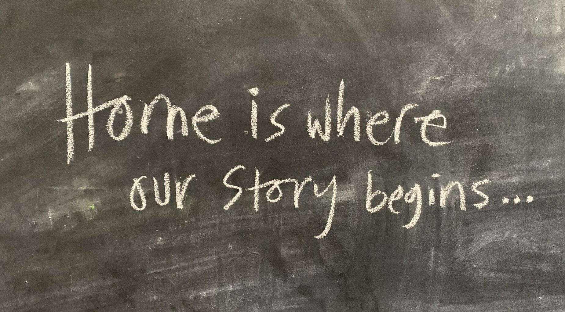 Chalkboard background with handwritten text in white chalk reads "Home is where our story begins..." conveying warmth and a sense of beginning.
