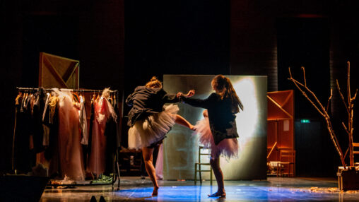 Alt text: Two dancers wearing dark jackets and pale tutus perform on a dimly lit stage. They move in a spotlight surrounded by set pieces including a clothing rail full of costumes, scattered shoes, chairs, and a bare tree-like prop in a box. The lighting creates a strong contrast between the dancers and the darker edges of the stage.
