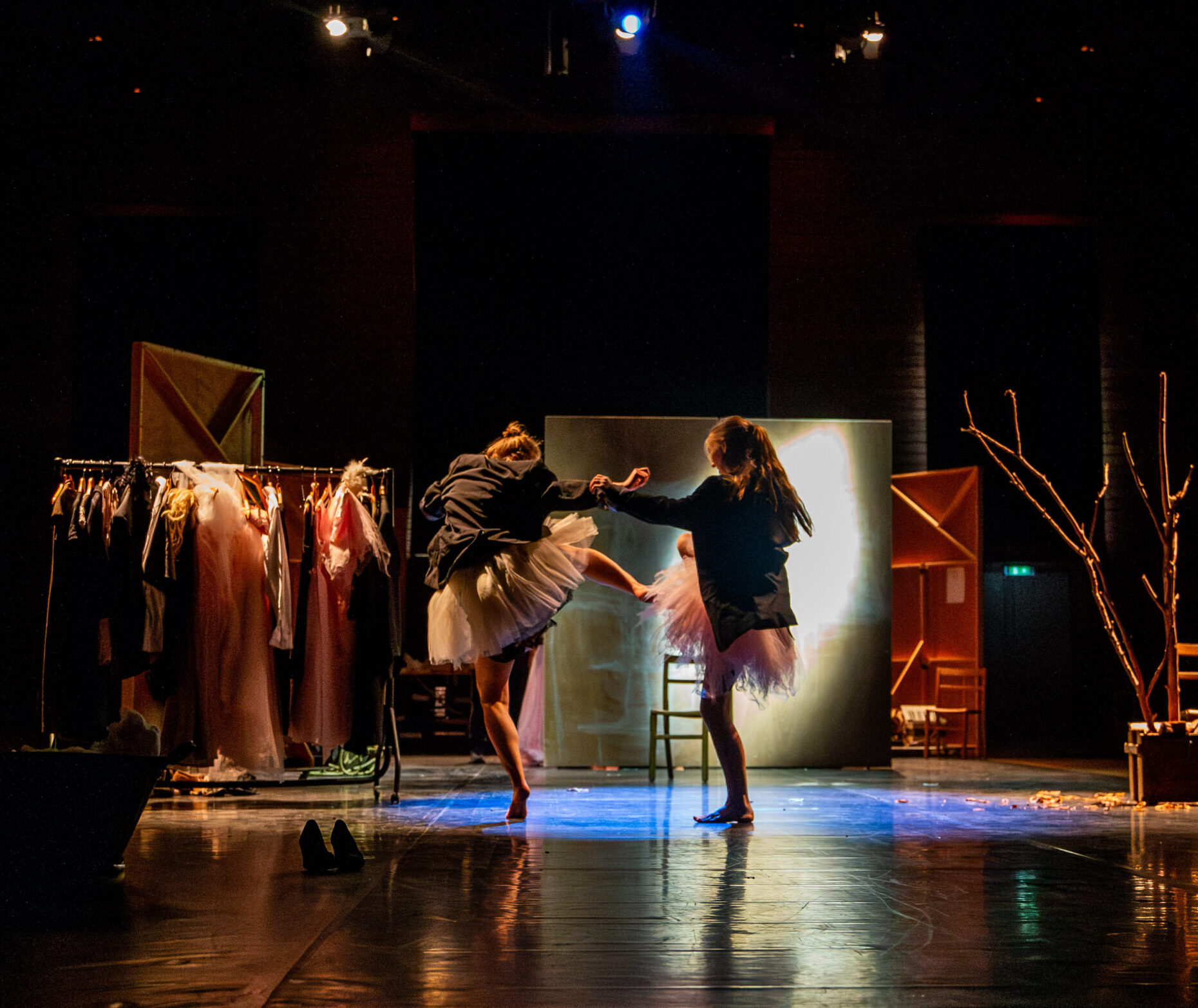 Alt text: Two dancers wearing dark jackets and pale tutus perform on a dimly lit stage. They move in a spotlight surrounded by set pieces including a clothing rail full of costumes, scattered shoes, chairs, and a bare tree-like prop in a box. The lighting creates a strong contrast between the dancers and the darker edges of the stage.